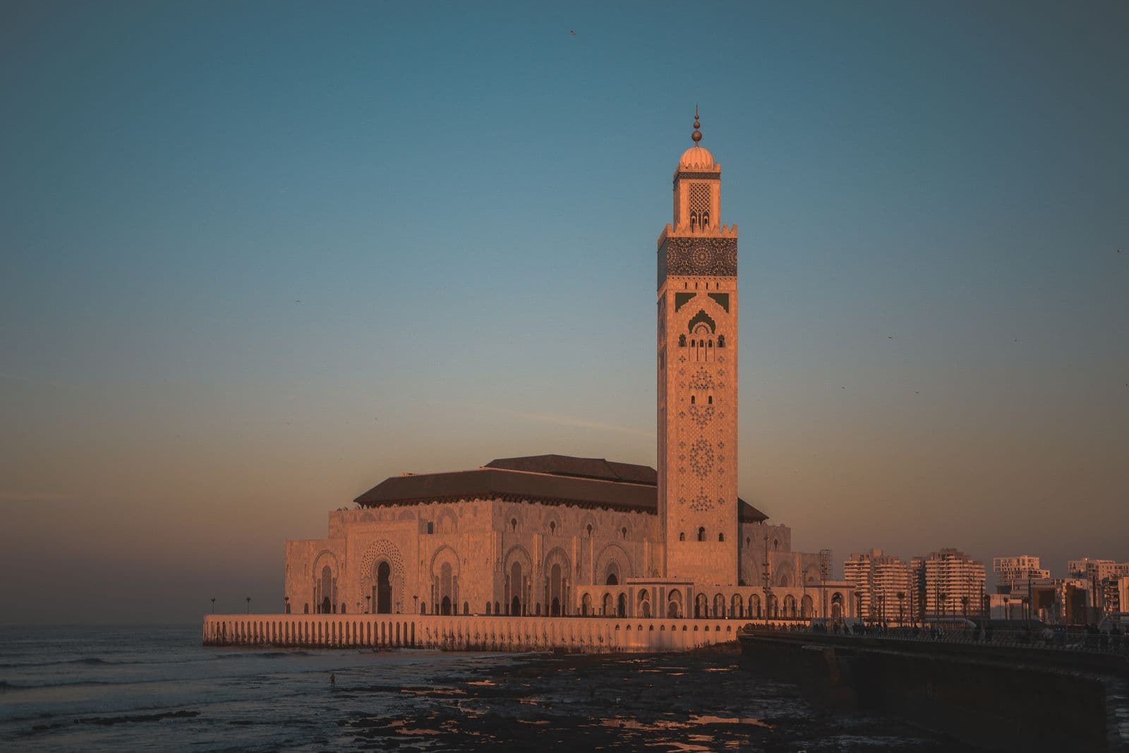 The Hassan II Mosque in Casablanca at sunset, its minaret rising above the sea wall with Atlantic surf breaking below