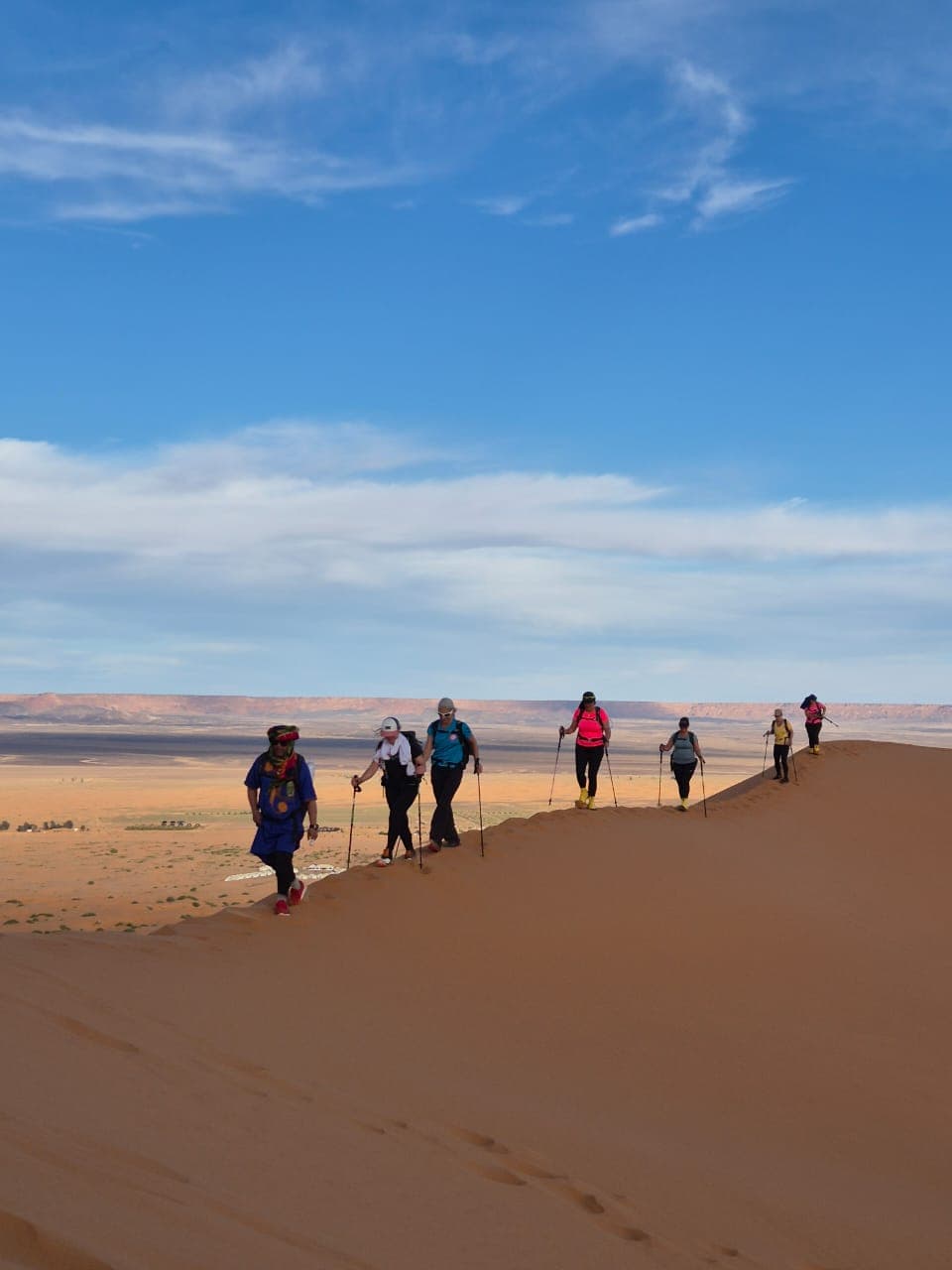 A small trekking group walking single file along a sunlit dune ridge, the stone hamada and a distant escarpment behind them