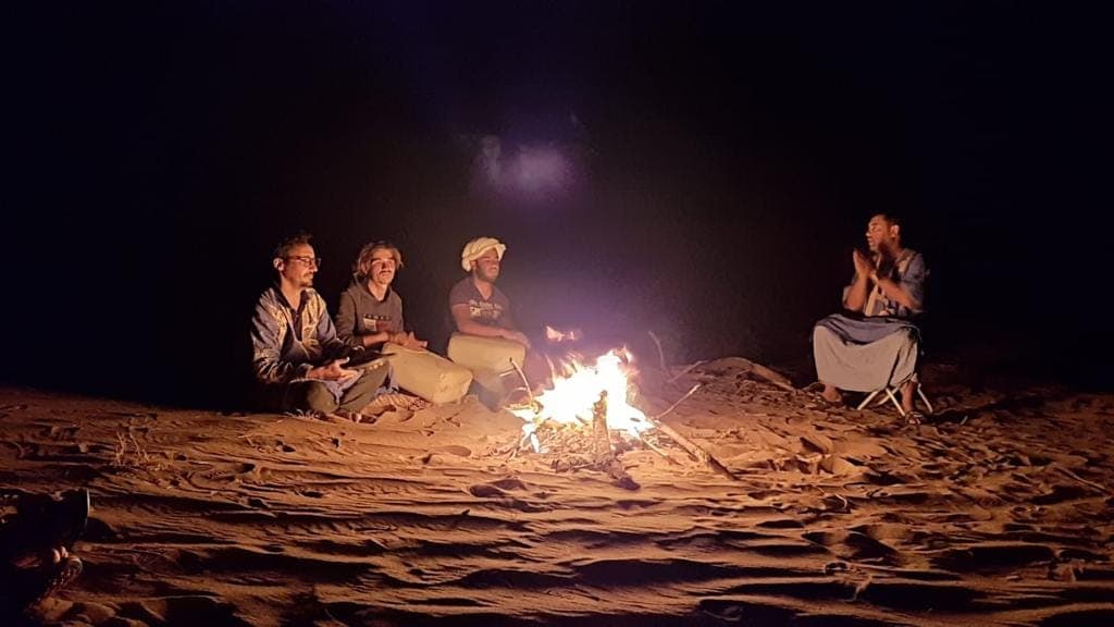 Four people sitting cross-legged on the sand around a small fire at night in the Sahara, one clapping a rhythm