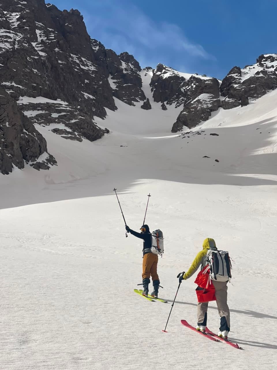 Two ski tourers skinning across a snow bowl below a rocky cirque in the High Atlas, one raising both poles