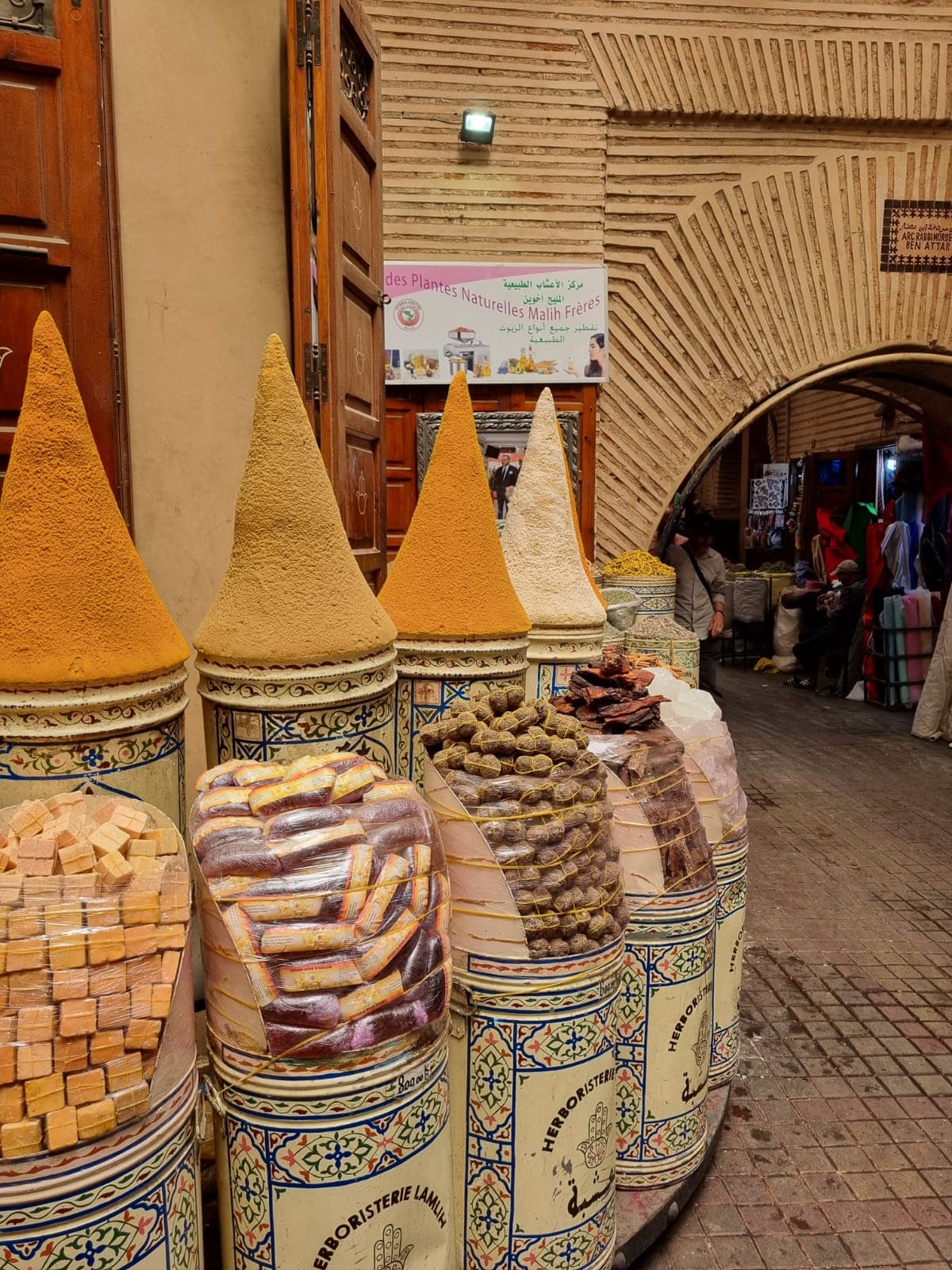 Cones of ground spices and stacked soap outside an herbalist's shop in a vaulted souk arcade, shoppers passing beyond