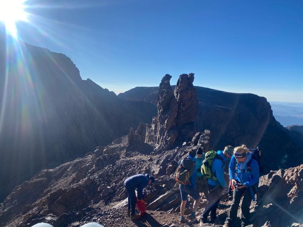 Trekkers in insulated jackets on the rocky summit ridge of Toubkal at first light, jagged pinnacles and the final plateau behind them