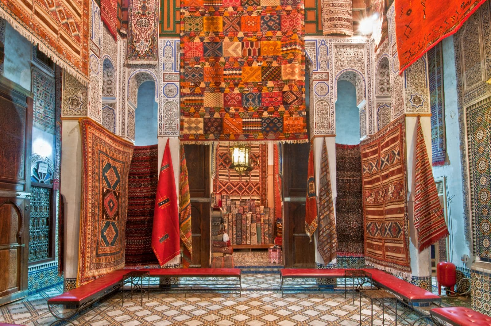Amazigh flatweaves and pile carpets hung floor to ceiling in a Fez carpet fondouk, zellij and carved plaster behind