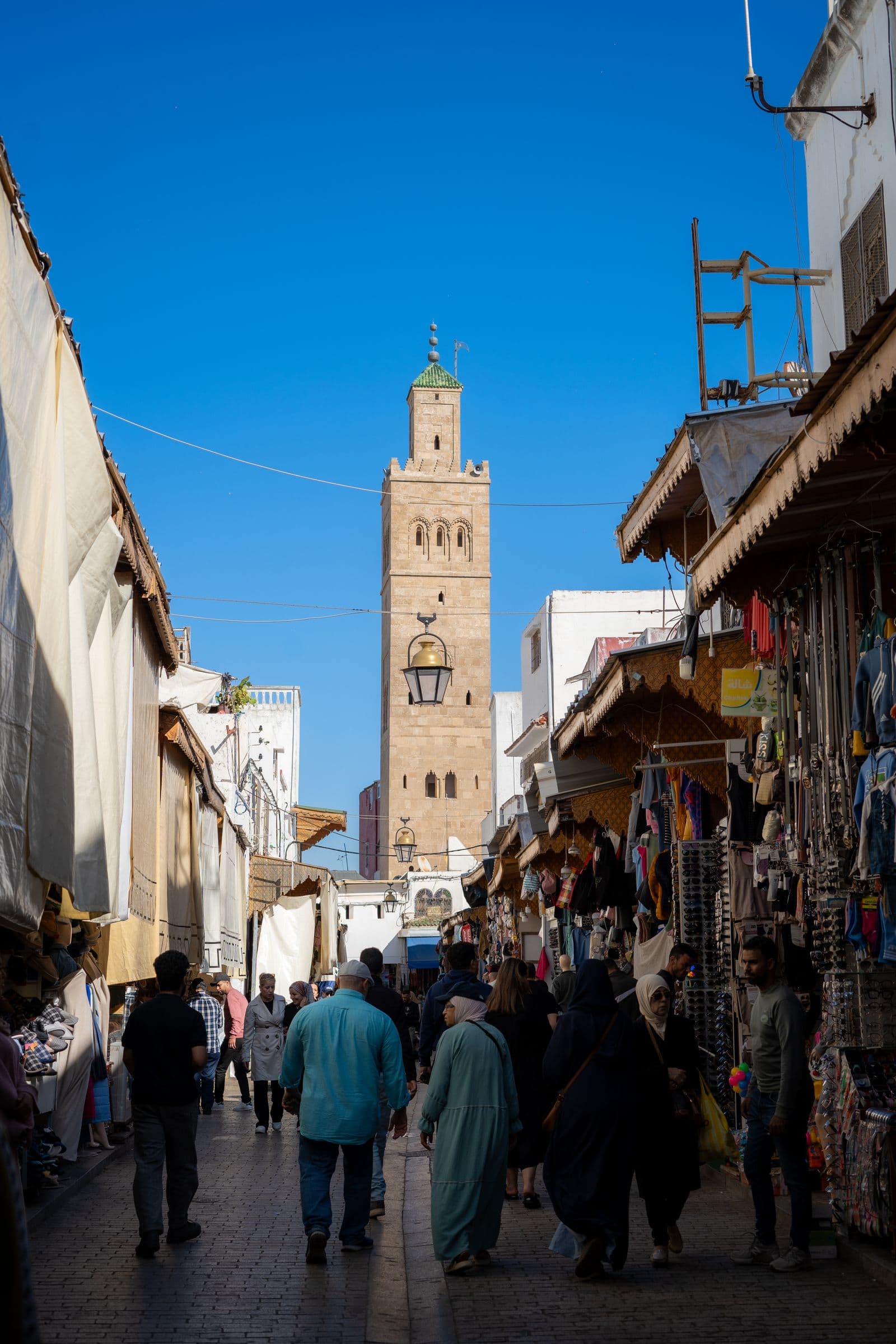 A bustling Marrakech souk lane with a historic minaret rising behind the rooftops