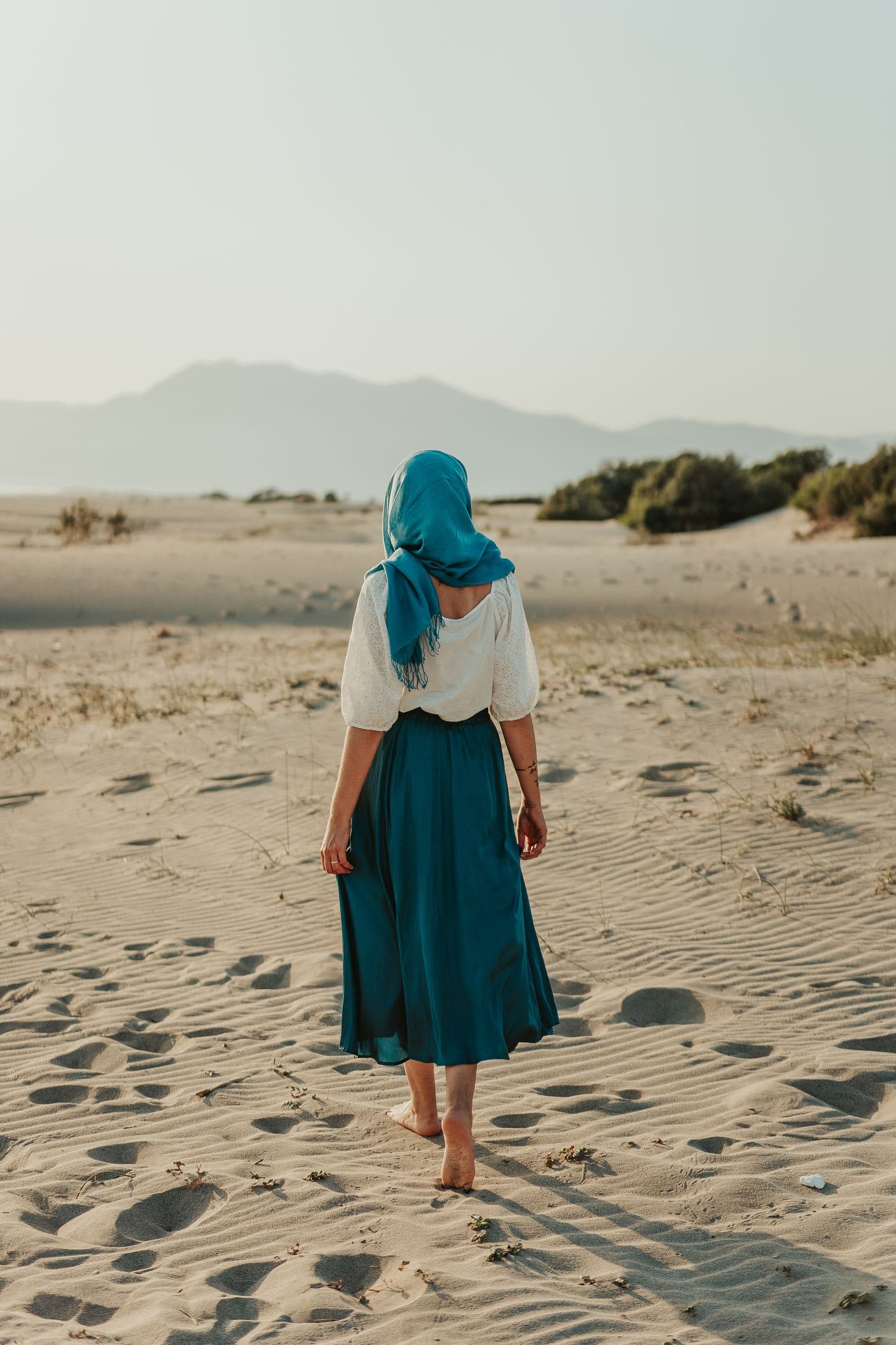 A woman travelling alone, headscarf and long skirt, walking away across open sand