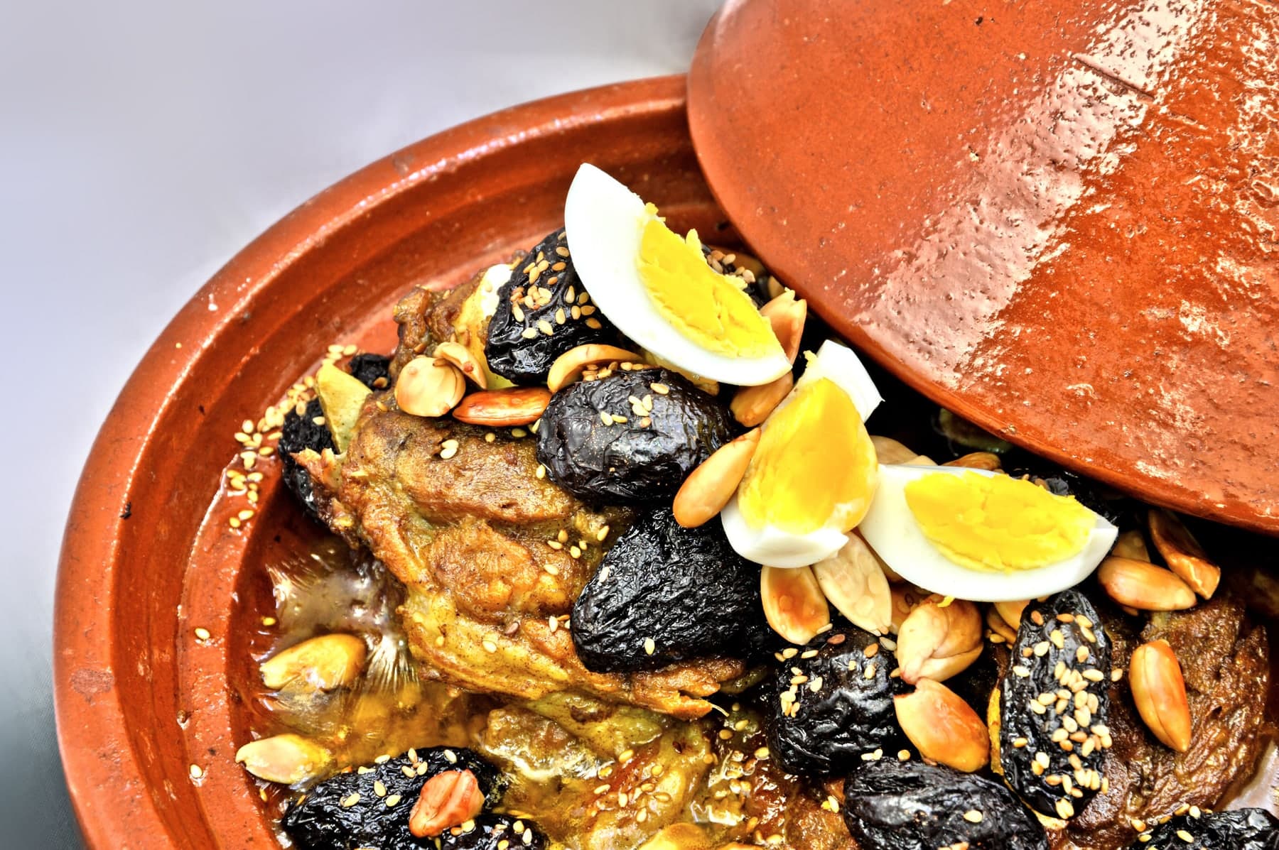 A clay tagine with the lid lifted, beef with prunes, toasted almonds, sesame seeds and hard-boiled egg inside